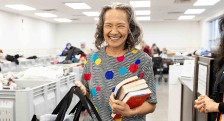 Smiling person holding books and shopping bags at a Women in Need Society community resource center, with volunteers and donation bins visible in the background