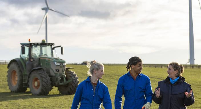 Three young adults in conversation in a farm field. A tractor and wind turbine are in the background.